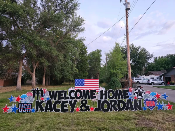 A yard sign that says "WELCOME HOME KACEY & JORDAN" in black letters. A graphic of a large American flag stands behind the phrase. Colorful graphics of balloons and stars surround the sign.
