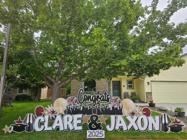 A yard sign that says "Congrats Clare & Jaxon" in black and white letters. Colorful graphics of balloons and stars surround the sign.