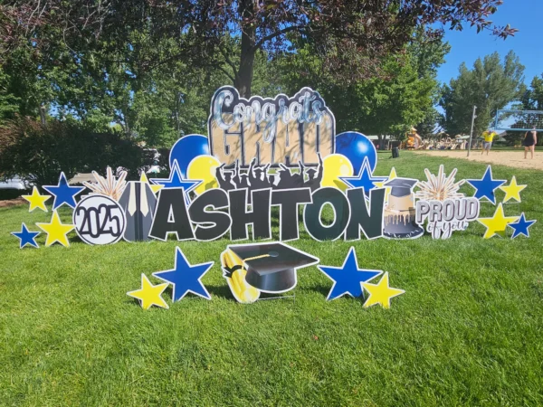 A large yard sign in front of a house that says "Congrats Grad Ashton" in colorful letters. Colorful graphics surround the sign.