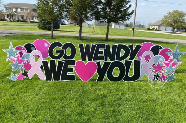 A yard sign that says "GO WENDY WE ♥ YOU" in black letters. Colorful graphics of balloons and stars surround the sign.