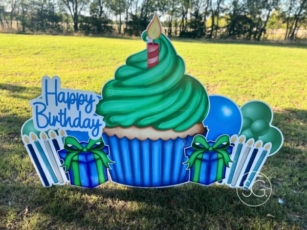 A yard sign in grassy field that says "Happy Birthday". The phrase is next to a large graphic of a cupcake and surrounded by colorful graphics.