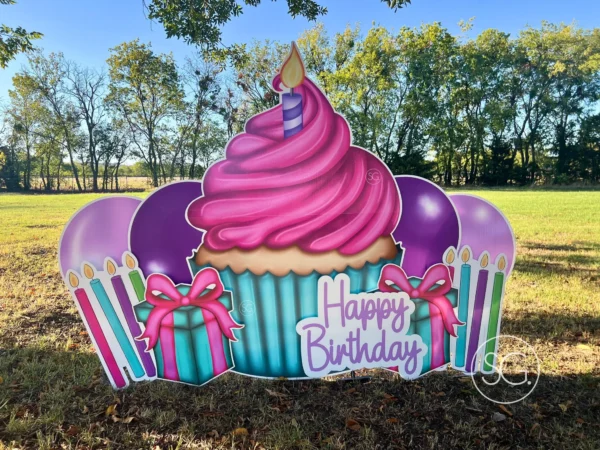 A yard sign in grassy field that says "Happy Birthday". The phrase is next to a large graphic of a cupcake and surrounded by colorful graphics.