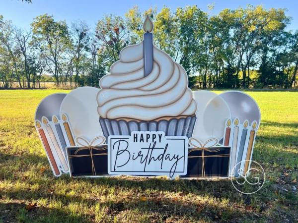 A yard sign in grassy field that says "Happy Birthday". The phrase is next to a large graphic of a cupcake and surrounded by colorful graphics.