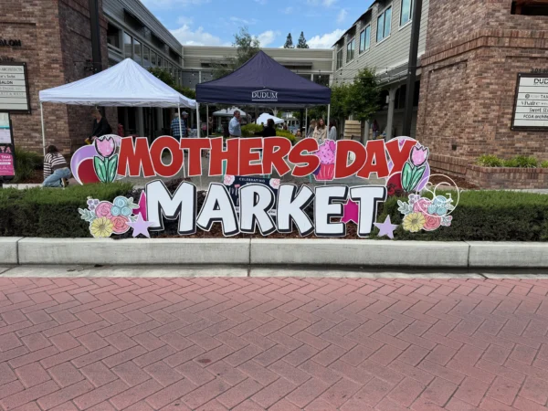 A yard sign in front of building that says "MOTHERS DAY MARKET" in red and white letters. Colorful graphics of flowers surround the sign.