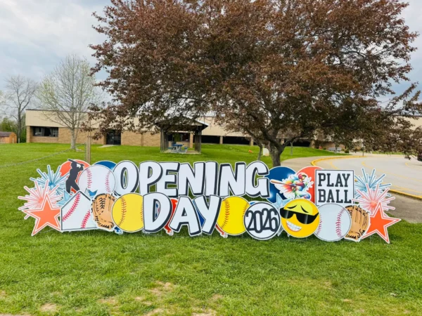A yard sign in front of building and a tree that says "OPENING DAY" in white letters. Colorful graphics surround the sign.