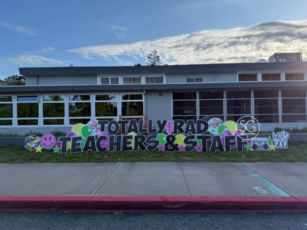 A yard sign in front of building that says "TOTALLY RAD TEACHERS & STAFF" in black letters. Colorful graphics surround the sign.