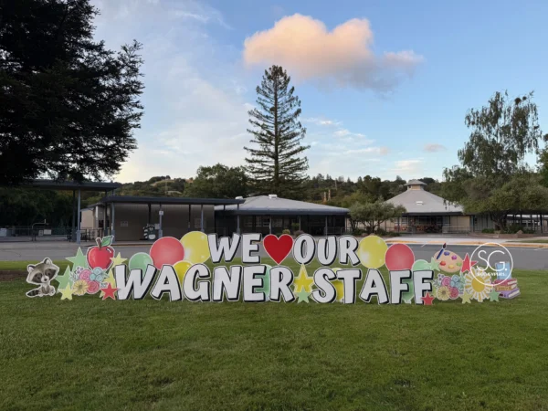 A yard sign in front of buildings that says "WE ♥ OUR WAGNER STAFF" in white letters. Colorful graphics surround the sign.