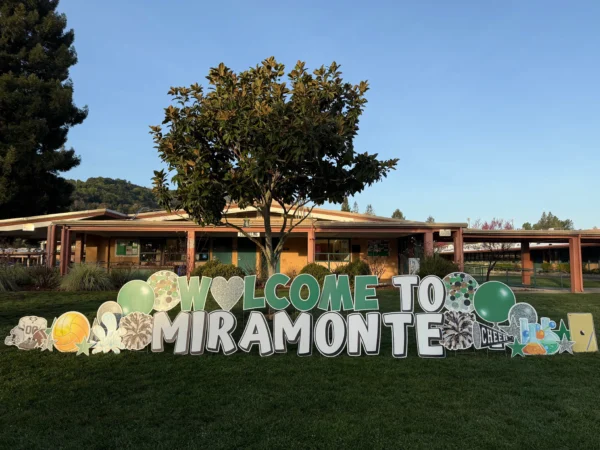 A yard sign in front of building that says "WELCOME TO MIRAMONTE" in green and white letters. Colorful graphics surround the sign.
