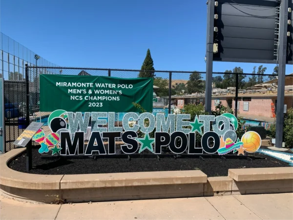 A yard sign in front of a swimming pool that says "WELCOME TO MATS POLO" in silver and black letters. Colorful graphics surround the sign.
