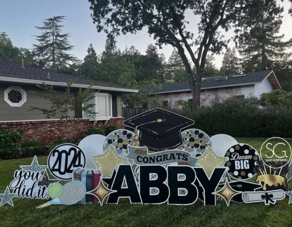 A large yard sign in front of a house that says "Congrats Abby" in black letters. Colorful graphics surround the sign.