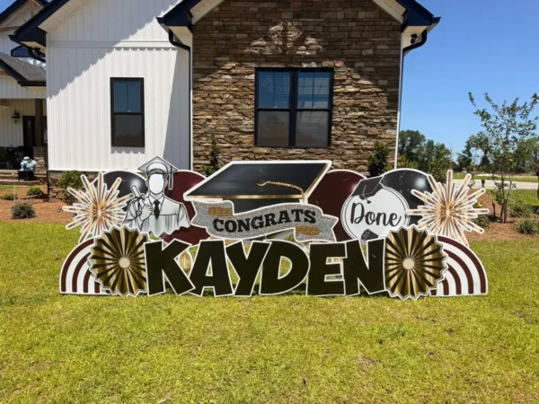A large yard sign in front of a house that says "Congrats Kayden" in black letters. Colorful graphics surround the sign.
