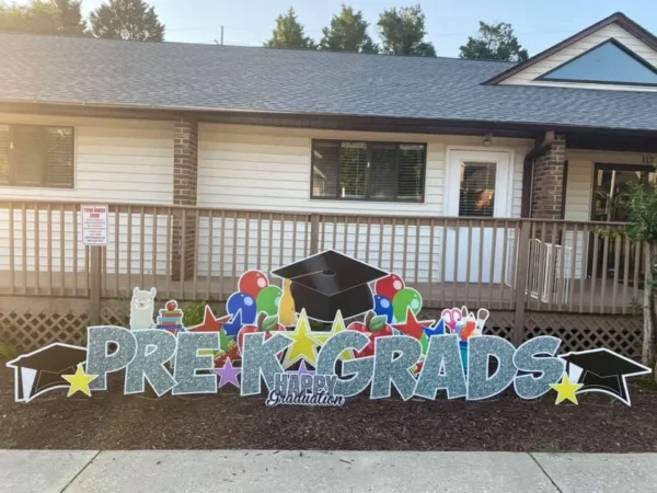 A large yard sign in front of a house that says "Pre K Grads Happy Graduation" in silver letters. Colorful graphics surround the sign.
