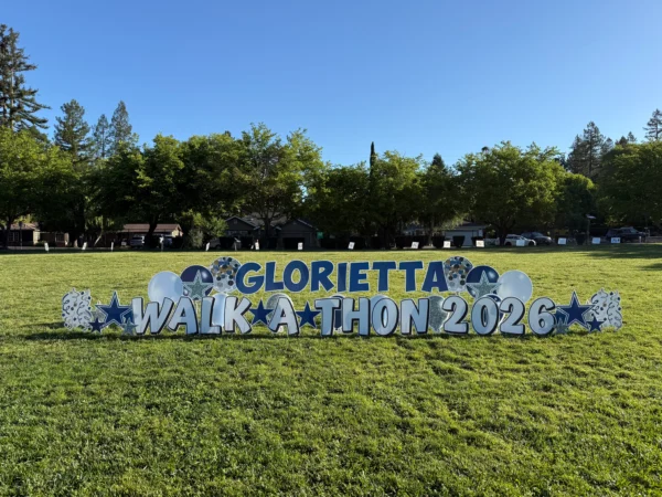 A yard sign in a grassy field that says "GLORIETTA WALKATHON 2026" in white and blue letters. Colorful graphics surround the sign.