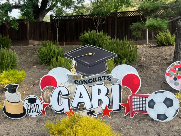 A large yard sign in front of a house with an image of a graduation cap that says "Congrats Abby" in black and white letters. Colorful graphics surround the sign.