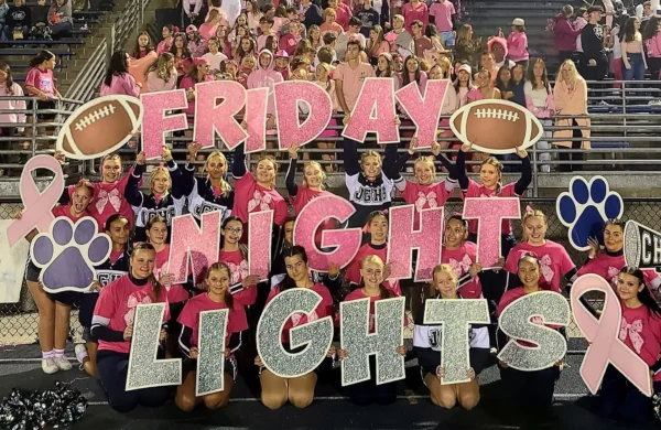 Kids at a sports event in a crowd holding up signs and letters that spell out "FRIDAY NIGHT LIGHTS" in pink and silver letters.
