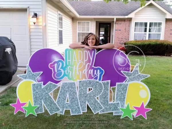 A yard sign in front of a house that says "Happy Birthday Karli" in colorful letters. The phrase is surrounded by colorful graphics. A girl stands behind the sign smiling.