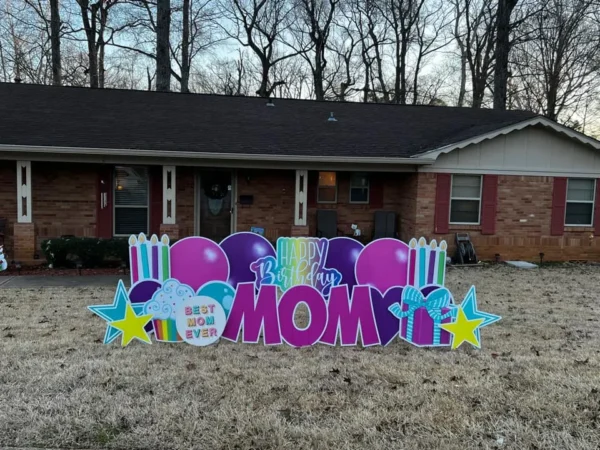 A yard sign in front of a house and tree that says "Happy Birthday Mom" in colorful letters. The phrase is surrounded by colorful graphics.