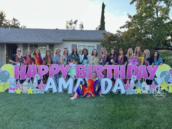 A yard sign in front of a house that says "HAPPY BIRTHDAY AMANDA" in pink and white letters. Colorful graphics surround the letters and multiple women stand behind the sign and one women sits in front of the sign.