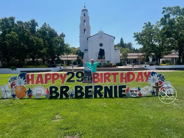 A yard sign in front of a church that says "HAPPY 29th BIRTHDAY BR. BERNIE" in red and black letters. Colorful graphics surround the letters and a man stands behind the sign with his arms raised.