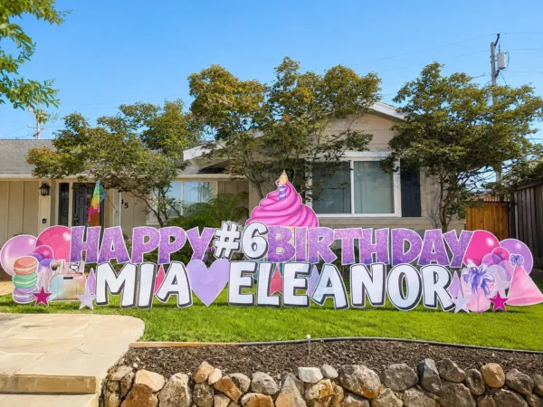 A yard sign in front of a house that says "HAPPY #6 BIRTHDAY MIA ELEANOR" in purple and white letters. A large cupcake sits behind the sign with colorful graphics around the letters.