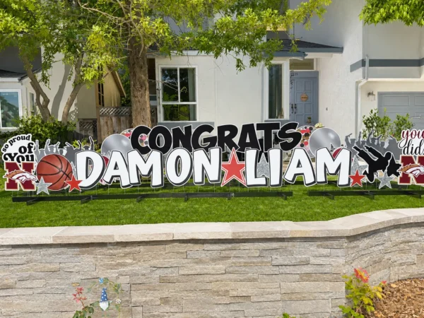 A large yard sign in front of a house that says "Congrats Damon Liam" in black and white letters. Colorful graphics surround the sign.