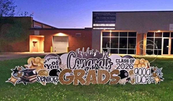 A large yard sign in front of a house that says "Congrats Grads" in white and gold letters. Colorful graphics surround the sign.