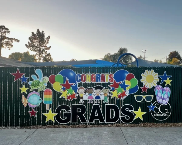 A large sign attached to a fence that says "Congrats Grads" in colorful letters. Colorful graphics surround the sign.