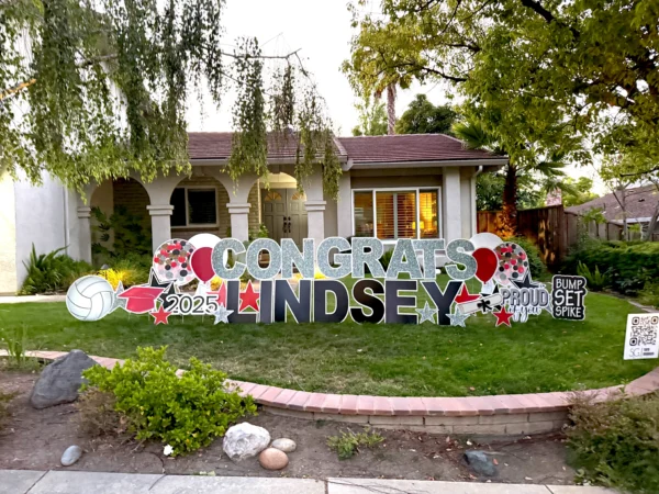 A large yard sign in front of a house that says "Congrats Lindsey" in silver and black letters. Colorful graphics surround the sign.