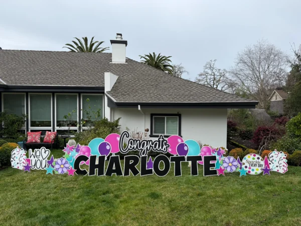A large yard sign in front of a house that says "Congrats Charlotte" in black letters. Colorful graphics surround the sign.