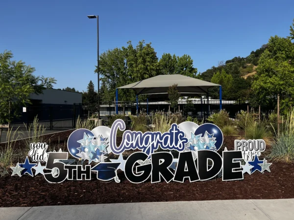 A large yard sign in front of a house that says "Congrats 5th Grade" in black and white letters. Colorful graphics surround the sign.